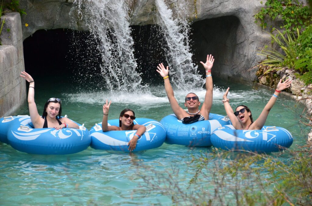 Four friends cheering on blue tubes in a lazy river beneath a jungle waterfall