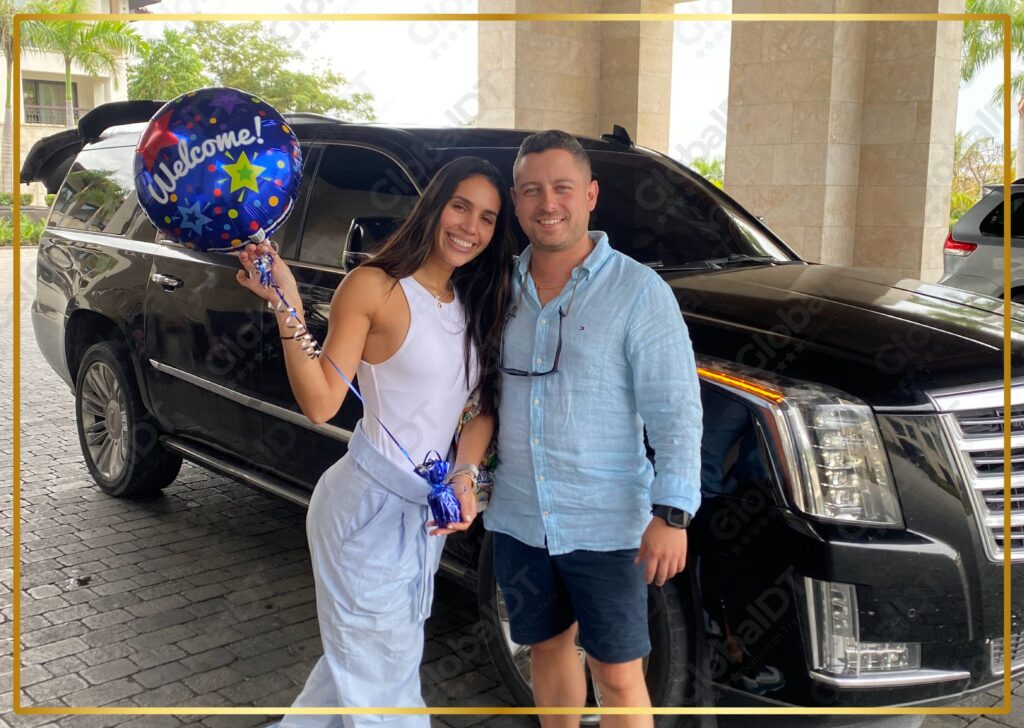 Couple posing with a Welcome balloon beside a black Cadillac Escalade at a resort entrance