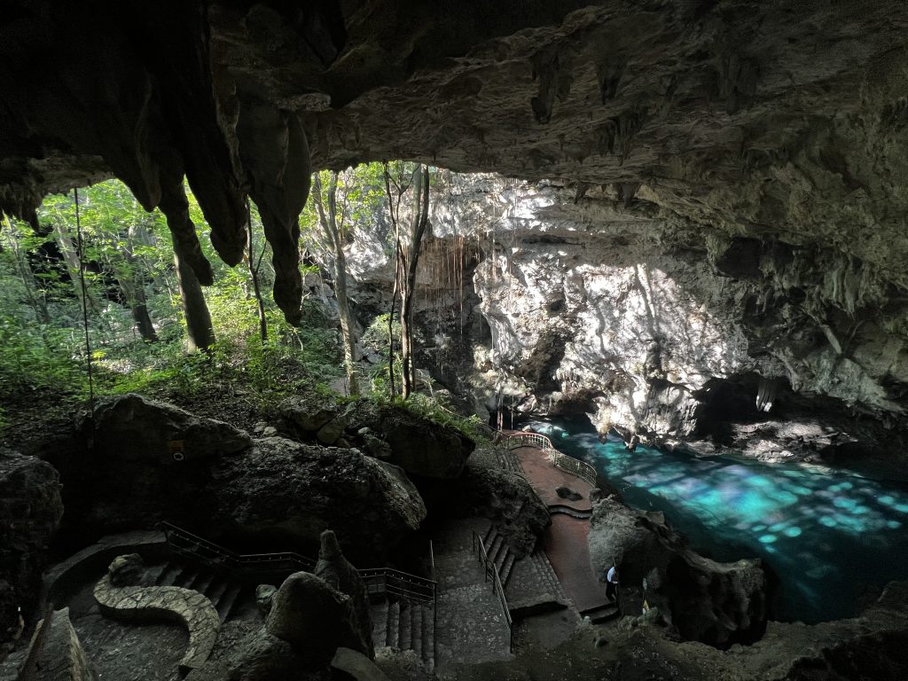 Cueva subterránea con agua turquesa, escalones de piedra y vegetación exuberante en la entrada