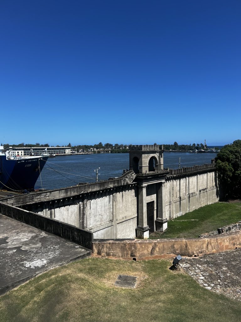 Fuerte histórico costero con vista a un puerto, un barco y cielo azul